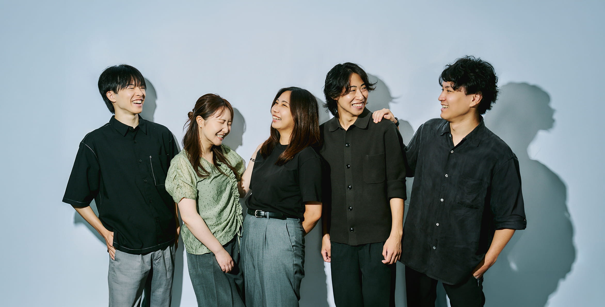 明るい背景の前で、笑顔で互いに顔を見合わせる私服姿の若手社員5人
